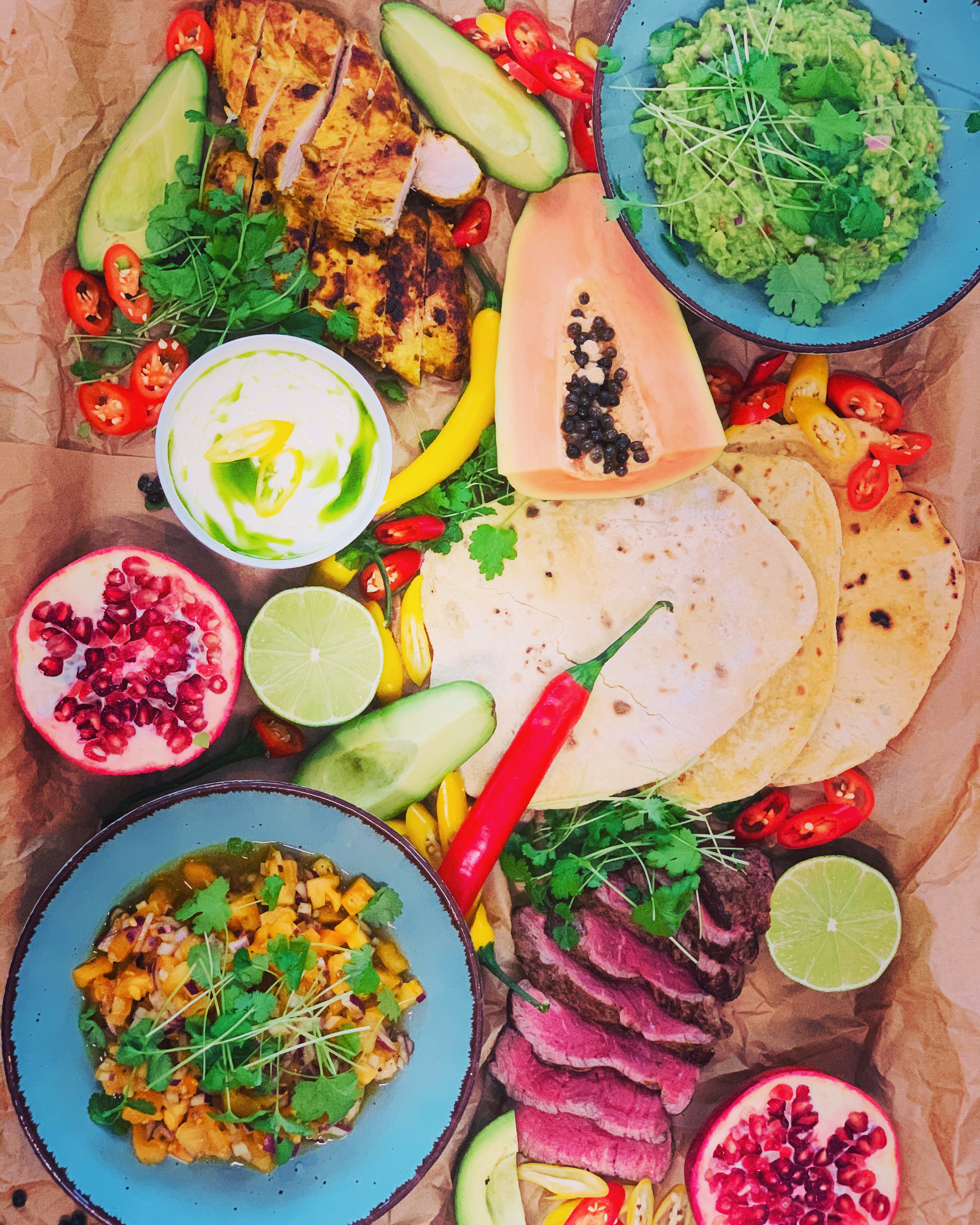 Widok z góry na aranżację talerzy i składników dania meksykańskiego, w tym grillowany kurczak, stek, guacamole, salsa mango, tortille, papaja, granat, limonka i chili, ułożonych na brązowym papierze.