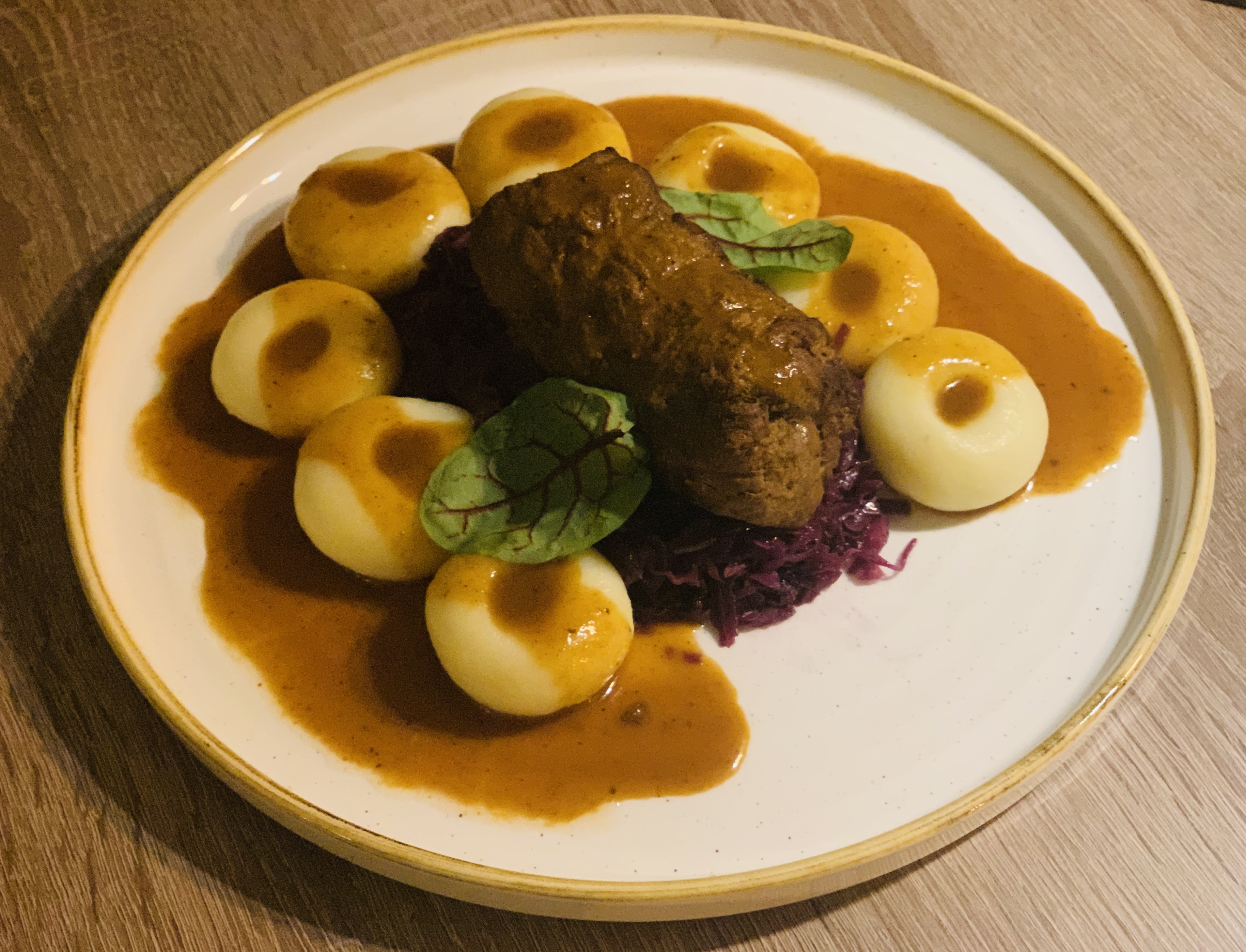 Gotowe danie: kluski śląskie z roladą mięsną, czerwoną kapustą i sosem na talerzu z drewnianym tłem.