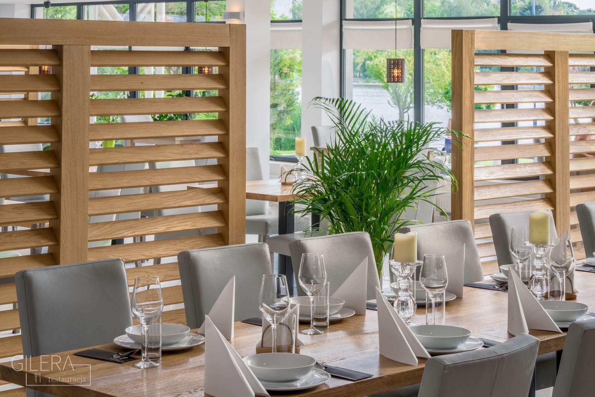 Elegancko nakryty stół w restauracji, z białymi obrusami, szklankami, świecami i zielonymi roślinami, widok na jezioro za oknem.