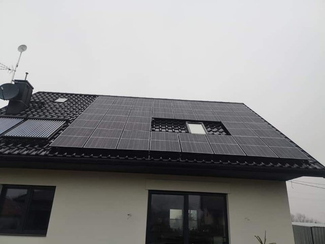 Czarny dach dwuspadowy z panelami słonecznymi i kolektorami słonecznymi, okno dachowe wkomponowane w panele, antena satelitarna na kominie, pochmurne niebo w tle.