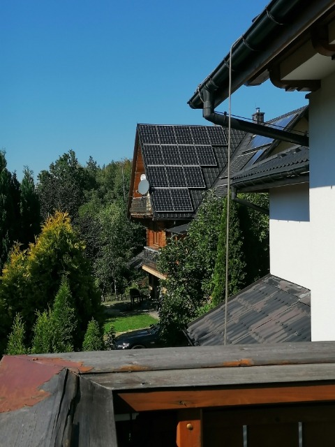 Widok na dom z panelami słonecznymi na dachu, z perspektywy innego budynku, w tle zieleń drzew i błękitne niebo.