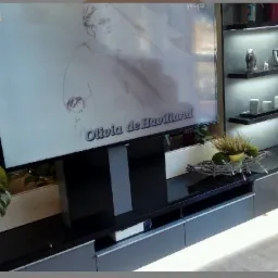 Ciemna szafka RTV z podświetleniem LED pod spodem, na niej telewizor z widoczną postacią Olivii de Havilland, obok regał z oświetleniem punktowym i dekoracjami.