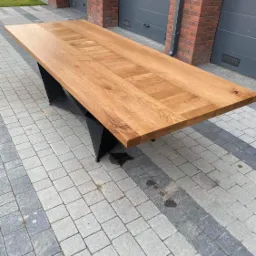 Drewniany stół na zewnątrz z blatem z jasnego drewna o wyraźnym usłojeniu i geometrycznym wzorze, osadzony na czarnej, metalowej podstawie o futurystycznym kształcie, na tle kostki brukowej...