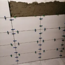 Białe płytki ścienne w trakcie montażu z użyciem krzyżyków dystansowych, widoczna warstwa kleju na górze ściany, częściowo ułożona podłoga z płytek.