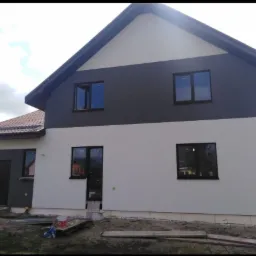 Dwukondygnacyjny dom po malowaniu elewacji, z szarą górną częścią i kremową dolną, widoczne puszki z farbą i narzędzia malarskie na trawniku.