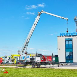 PPHU WINDEX Mirela Aszyk - Mobilny dźwig z wysięgnikiem podnosi element na dachu budynku z napisem WINDEX, w tle maszt telekomunikacyjny i inne maszyny na placu.