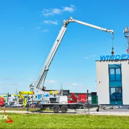Mobilny dźwig z wysięgnikiem podnosi element na dachu budynku z napisem WINDEX, w tle maszt telekomunikacyjny i inne maszyny na placu.