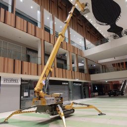 PPHU WINDEX Mirela Aszyk - Żółty podnośnik teleskopowy Ommelift 3150 ustawiony wewnątrz galerii handlowej, z wysięgnikiem skierowanym w stronę sufitu z dekoracją w kształcie wazonu.