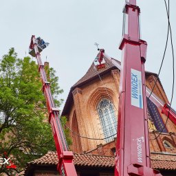 PPHU WINDEX Mirela Aszyk - Dwa czerwone podnośniki teleskopowe firmy Windex pracują na wysokości przy zabytkowej, ceglanej katedrze z wysokimi oknami i krzyżem na szczycie dachu.
