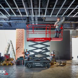 PPHU WINDEX Mirela Aszyk - Pracownik na czerwonym podnośniku nożycowym marki Windex, montujący instalację pod sufitem w surowym wnętrzu, widoczne narzędzia i materiały budowlane, obok drabiny i skrzynki narzędziowe.