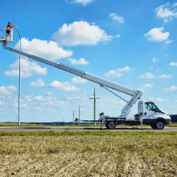 Biały pojazd z wysięgnikiem i koszem na tle błękitnego nieba z chmurami, pracownik w pomarańczowym ubraniu w koszu podczas pracy na wysokości, w tle pole i słupy energetyczne.