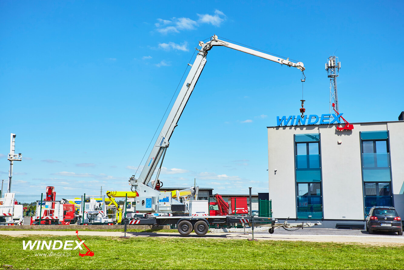 Mobilny dźwig z wysięgnikiem podnosi element na dachu budynku z napisem WINDEX, w tle maszt telekomunikacyjny i inne maszyny na placu.