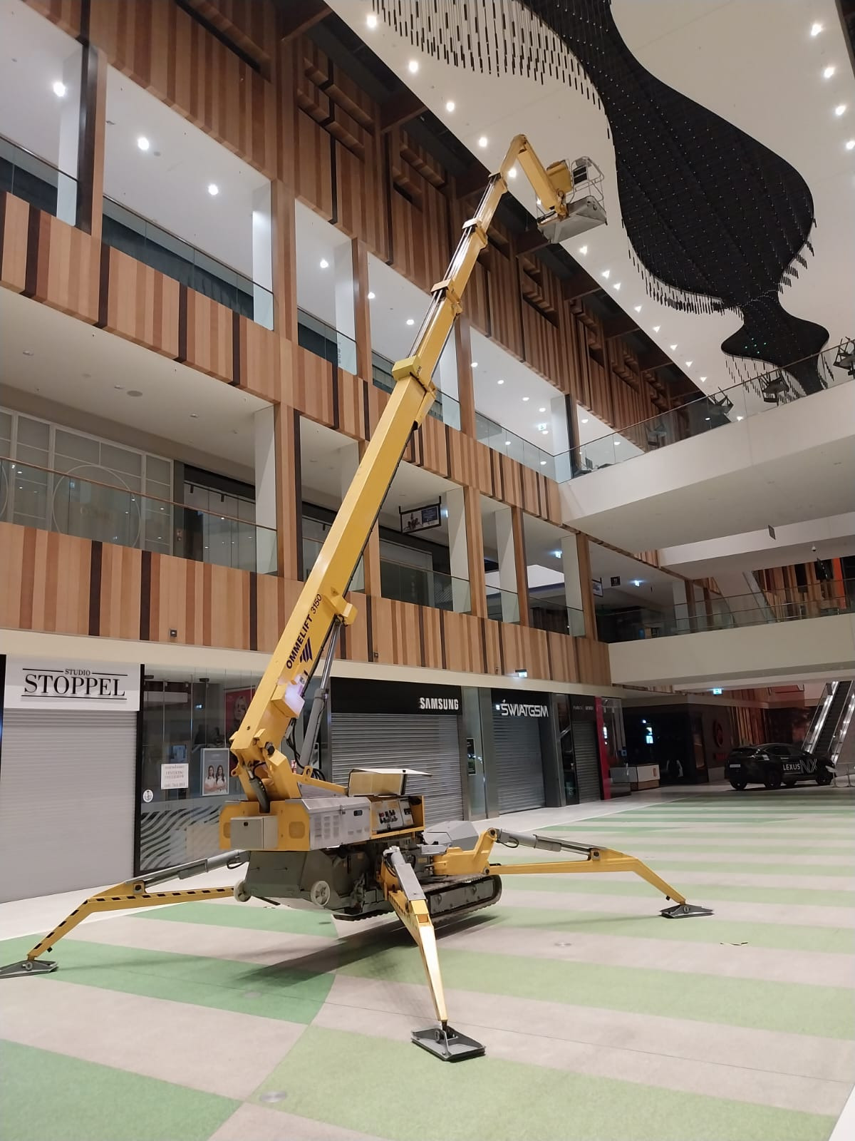 Żółty podnośnik teleskopowy Ommelift 3150 ustawiony wewnątrz galerii handlowej, z wysięgnikiem skierowanym w stronę sufitu z dekoracją w kształcie wazonu.