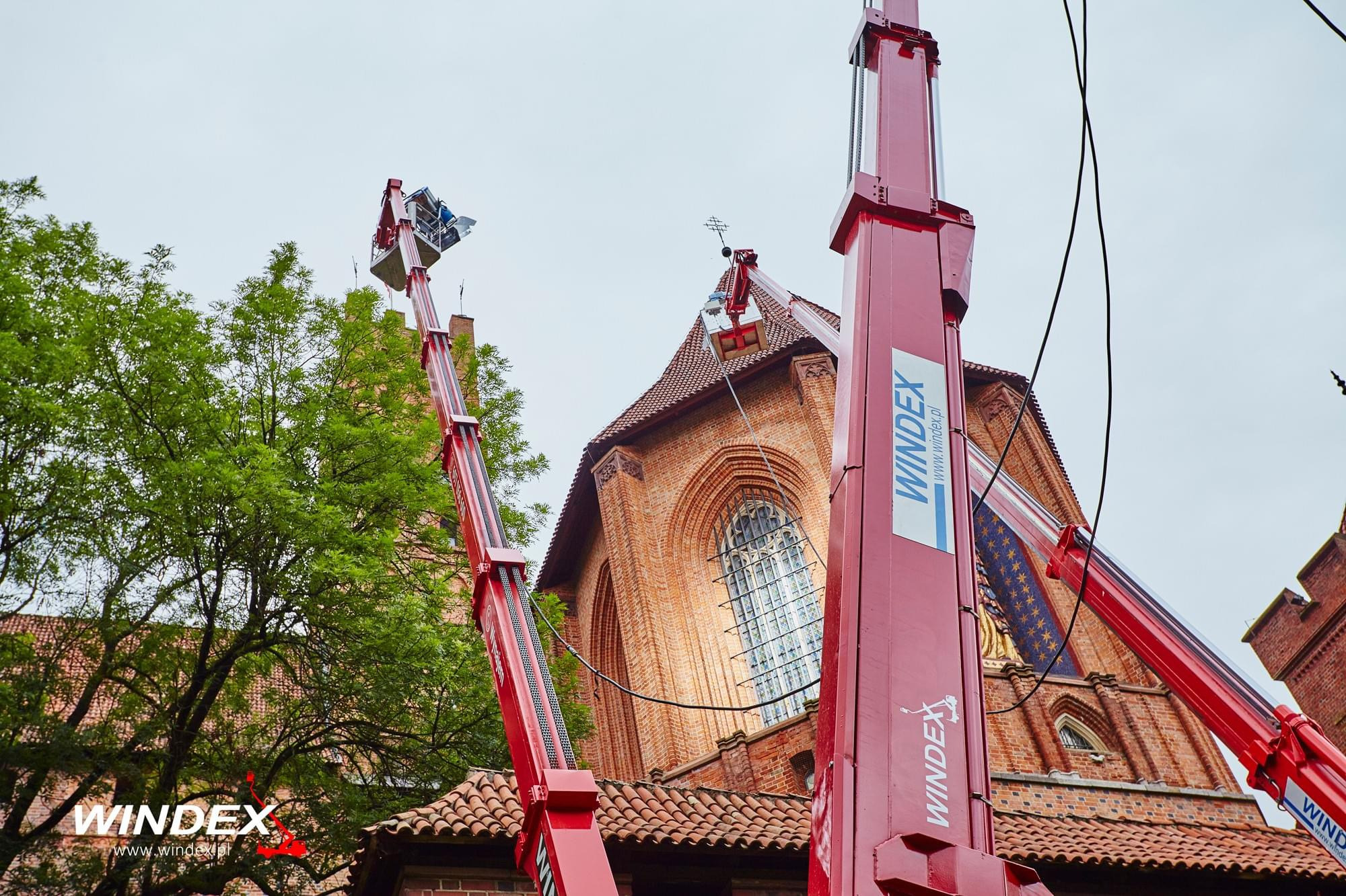Dwa czerwone podnośniki teleskopowe firmy Windex pracują na wysokości przy zabytkowej, ceglanej katedrze z wysokimi oknami i krzyżem na szczycie dachu.