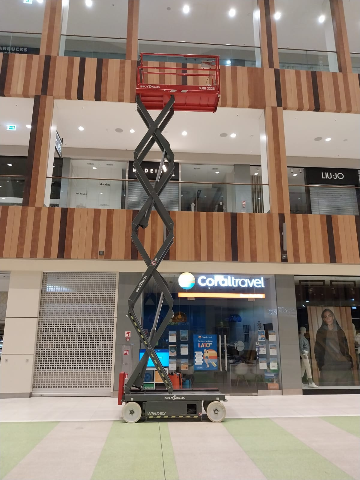 Czerwony podnośnik nożycowy Skyjack wewnątrz galerii handlowej, widoczne sklepy Coral Travel, MaxMara i Liu Jo w tle.