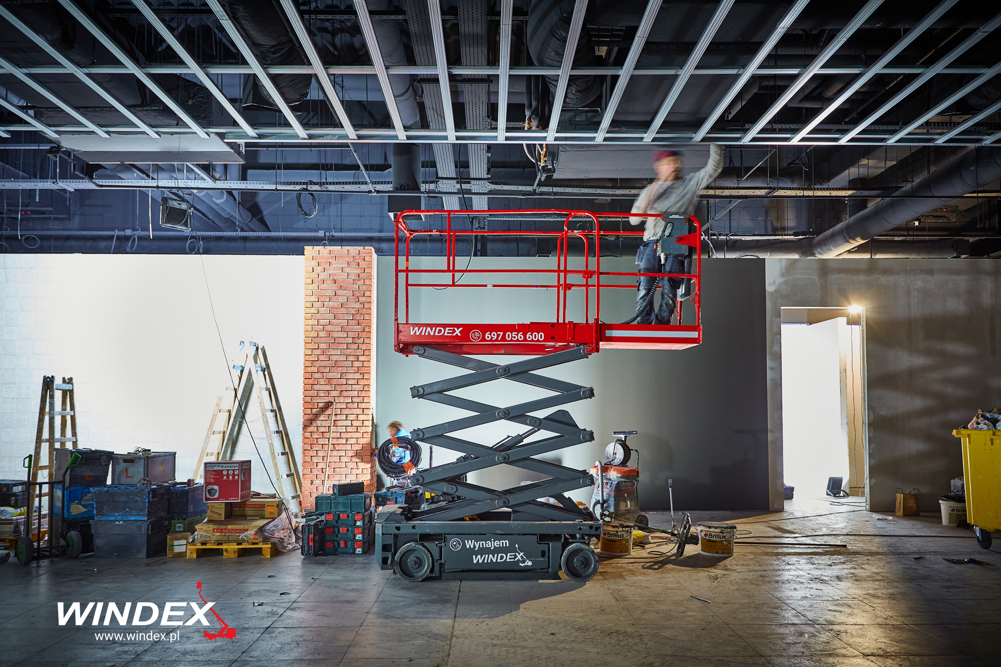 Pracownik na czerwonym podnośniku nożycowym marki Windex, montujący instalację pod sufitem w surowym wnętrzu, widoczne narzędzia i materiały budowlane, obok drabiny i skrzynki narzędziowe.