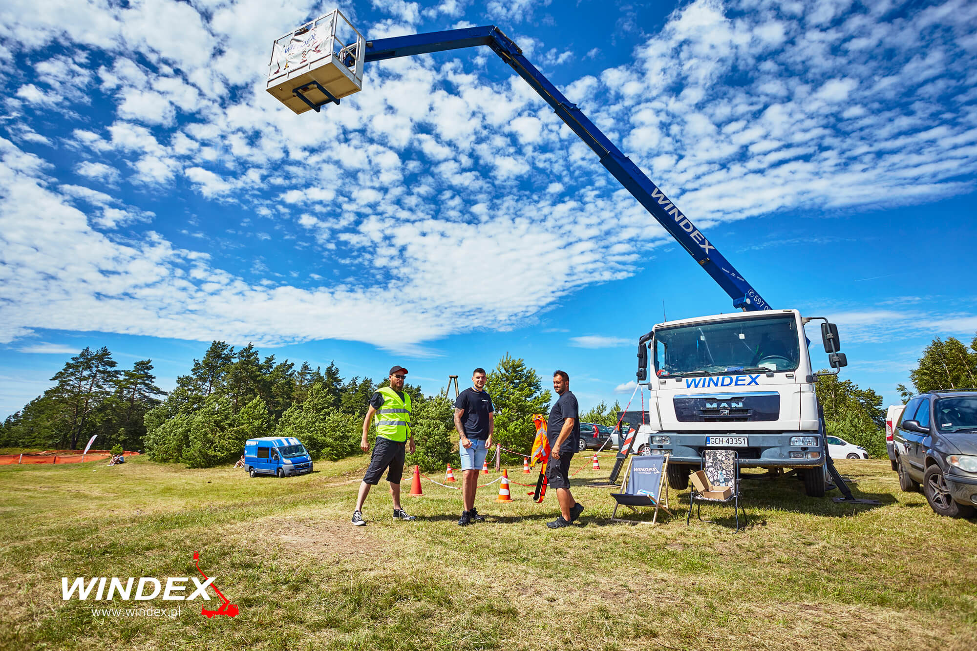 Trzech mężczyzn stoi przed podnośnikiem koszowym marki Windex na trawiastym terenie w słoneczny dzień. W tle widoczne lasy i błękitne niebo z obłokami.
