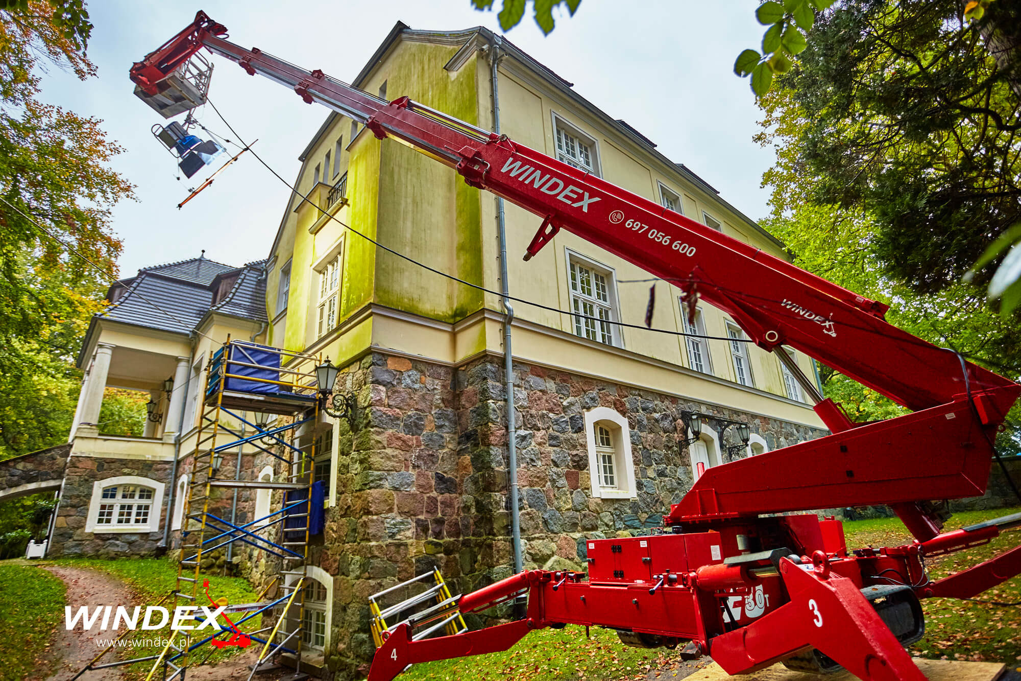 Czerwony podnośnik koszowy Windex z koszem i sprzętem oświetleniowym na tle odnowionej kamienicy z kamienną elewacją i rusztowaniami.