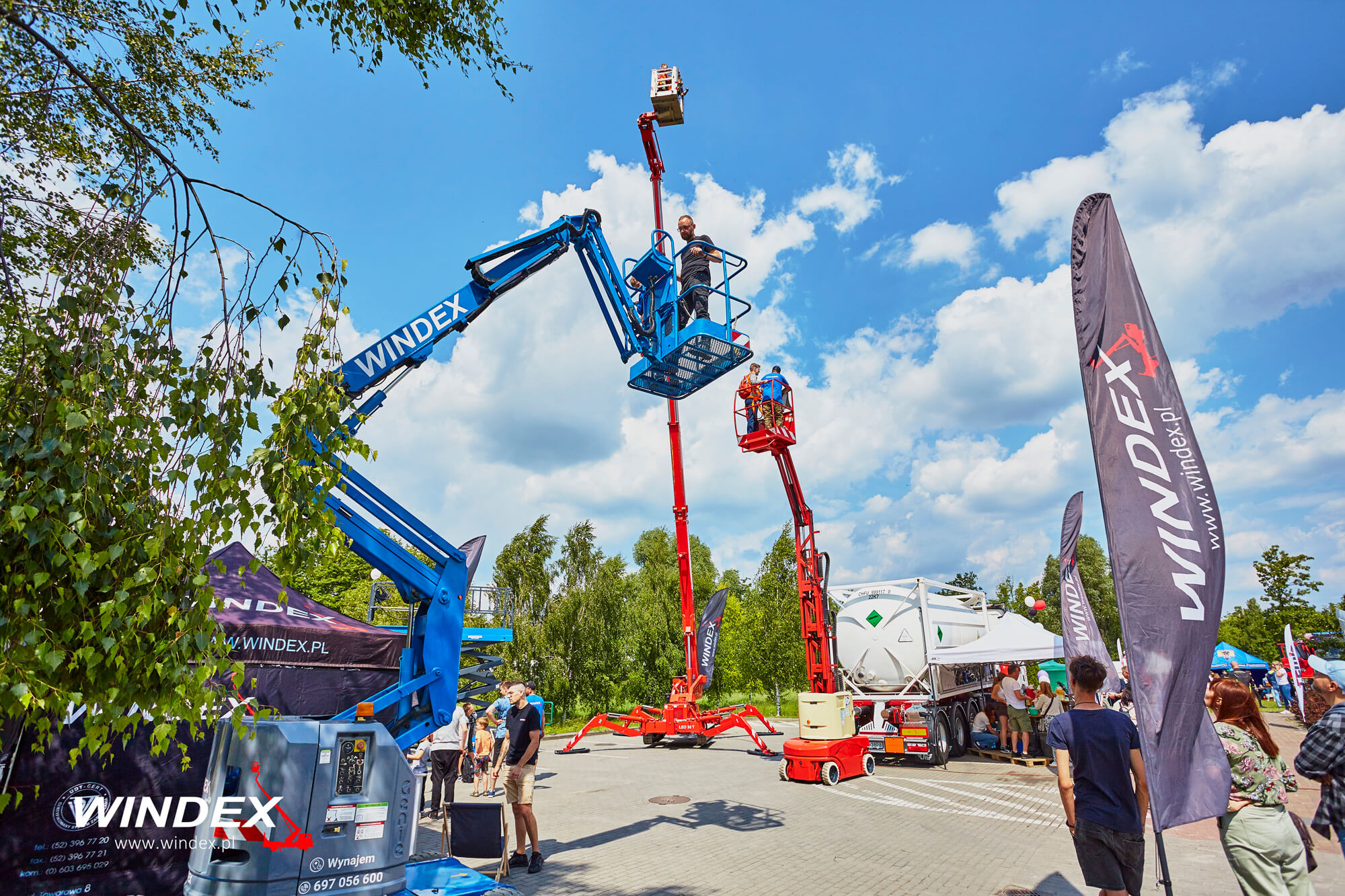 Dwa podnośniki przegubowe w kolorach niebieskim i czerwonym, z pracownikami w koszach, prezentowane na otwartej przestrzeni z drzewami w tle, obok zaparkowana cysterna, widoczne flagi reklamowe.