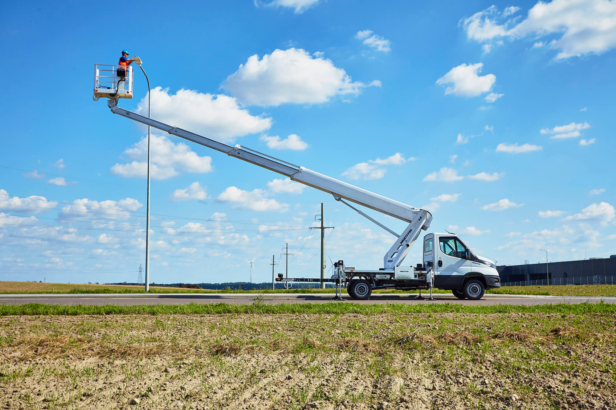 Biały pojazd z wysięgnikiem i koszem na tle błękitnego nieba z chmurami, pracownik w pomarańczowym ubraniu w koszu podczas pracy na wysokości, w tle pole i słupy energetyczne.