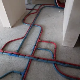 Rozłożone na surowym podłożu rury instalacji ogrzewania podłogowego, niebieskie i czerwone, przymocowane czarnymi uchwytami, w surowym stanie wykończenia pomieszczenia.