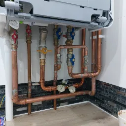 Montaż pieca gazowego z widocznymi miedzianymi rurami, zaworami z czerwonymi i niebieskimi pokrętłami, oraz fragmentem instalacji elektrycznej z białymi złączkami, na tle białej ściany z ozdobną...