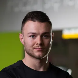 Portret młodego mężczyzny z krótką brodą, w czarnej koszulce, na tle ściany w odcieniach szarości i zieleni, z widocznym napisem w tle.