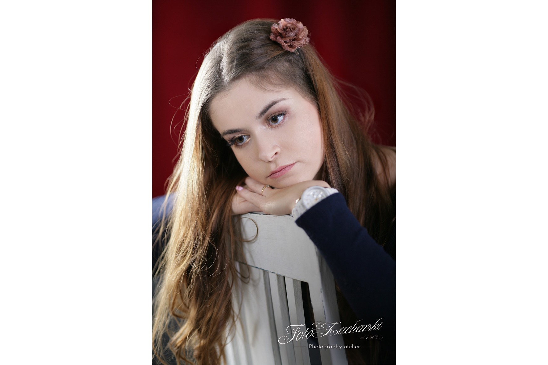 Portret młodej kobiety z brązowym kwiatem we włosach, opierającej się o białe krzesło na tle czerwonej ściany, w melancholijnej pozie.