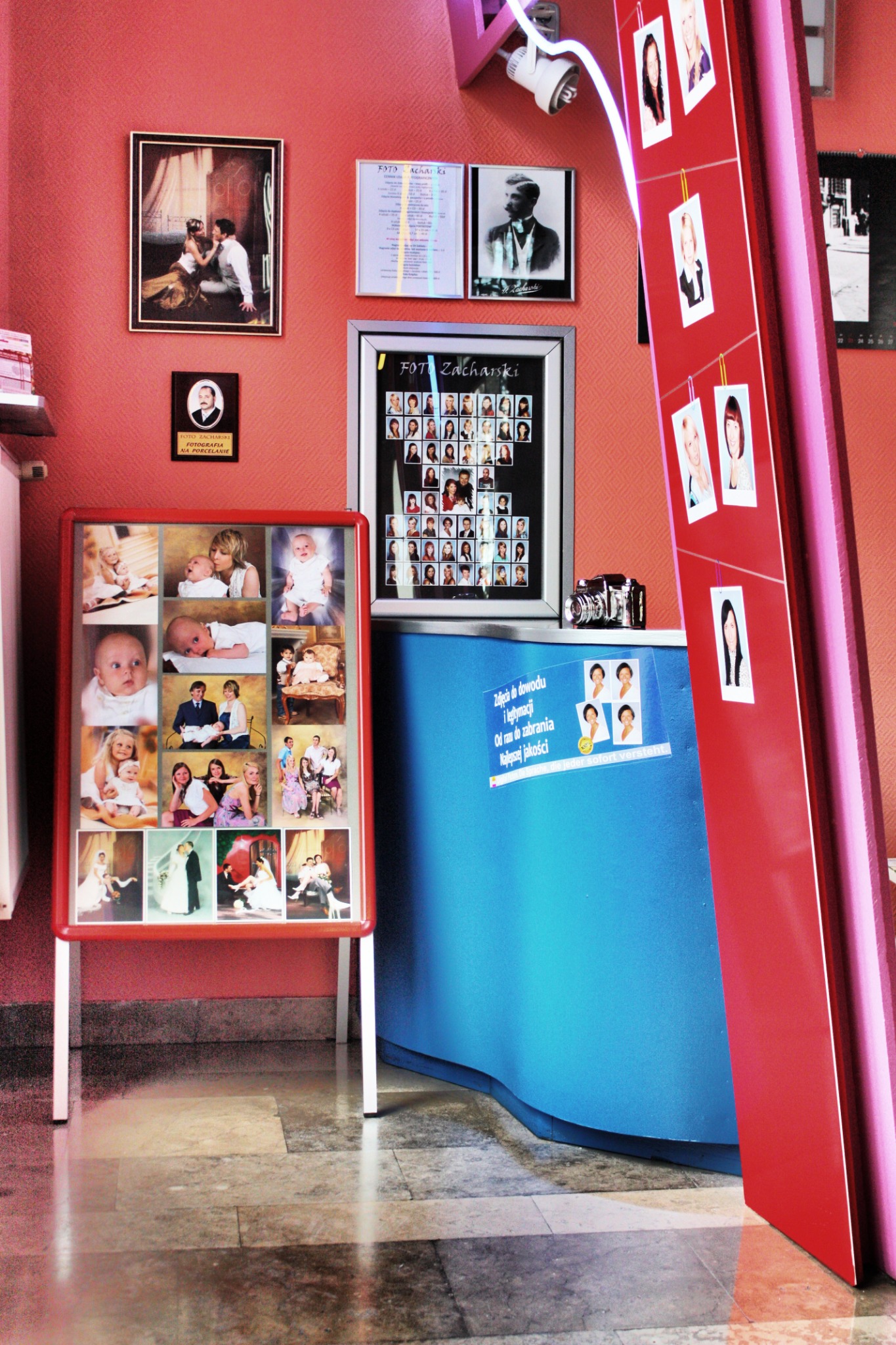 Wnętrze zakładu fotograficznego z ekspozycją zdjęć dzieci, noworodków i ślubnych par na stojaku, portrety na ścianie oraz aparat fotograficzny na ladzie.