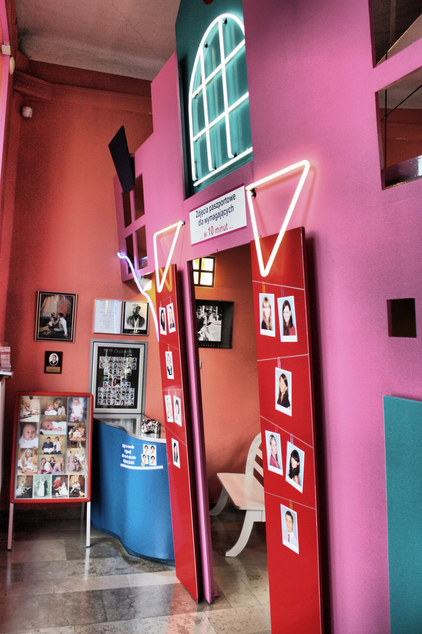 Wnętrze zakładu fotograficznego z neonowym oknem, czerwonymi panelami z przypiętymi zdjęciami portretowymi i ekspozycją zdjęć niemowląt, w pastelowej kolorystyce.