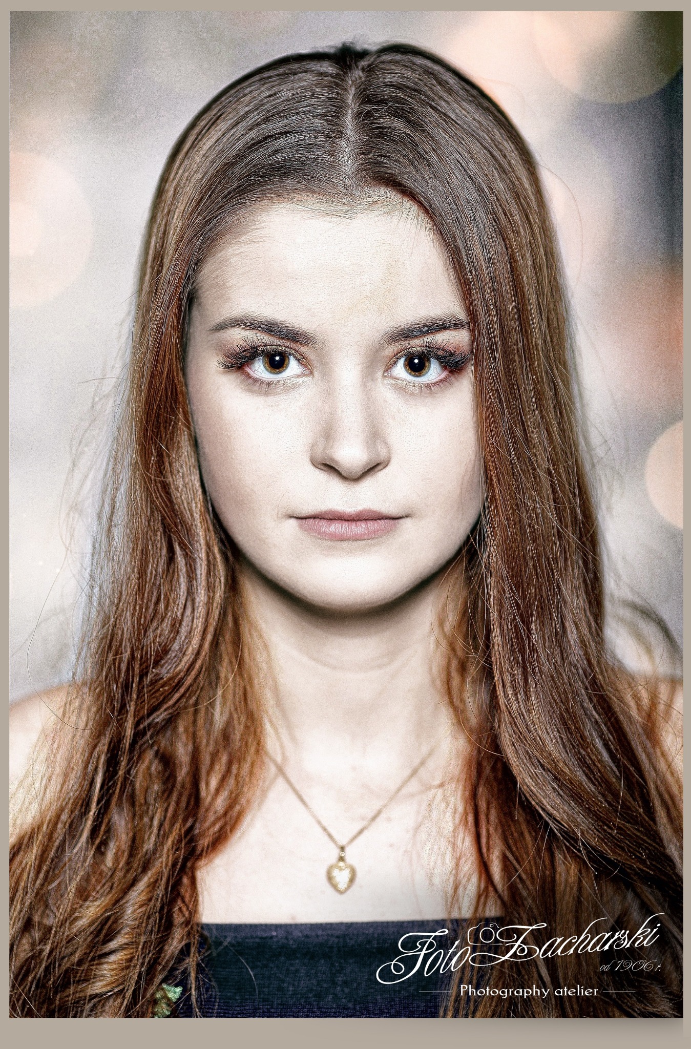 Portret młodej kobiety z długimi brązowymi włosami i delikatnym makijażem, naszyjnik z serduszkiem, na rozmytym tle z bokeh, w dolnym rogu sygnatura fotografa.