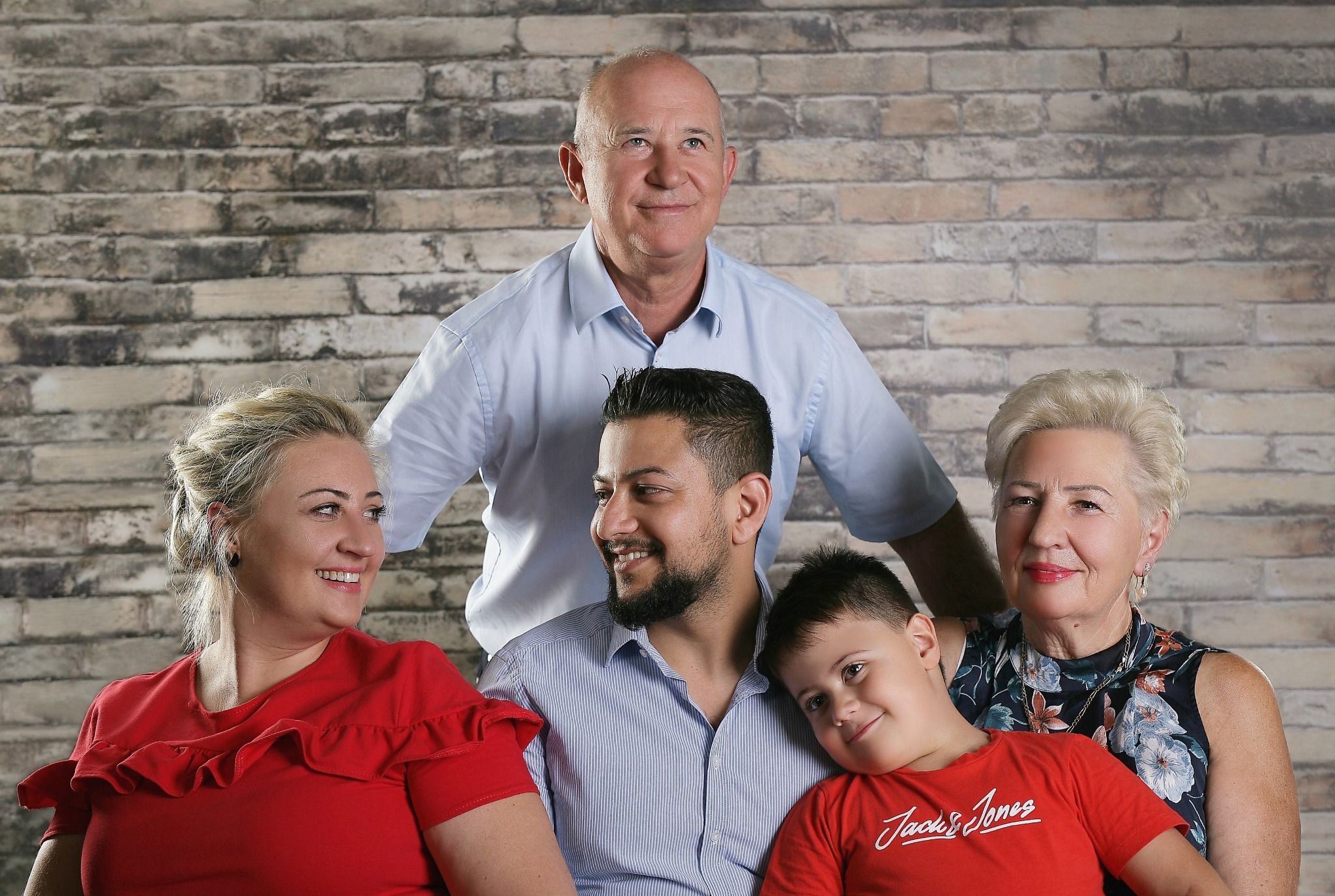 Pięcioosobowa rodzina pozuje do zdjęcia na tle ceglanej ściany. Dwie kobiety, dwóch mężczyzn i chłopiec w czerwonej koszulce Jack&Jones uśmiechają się.