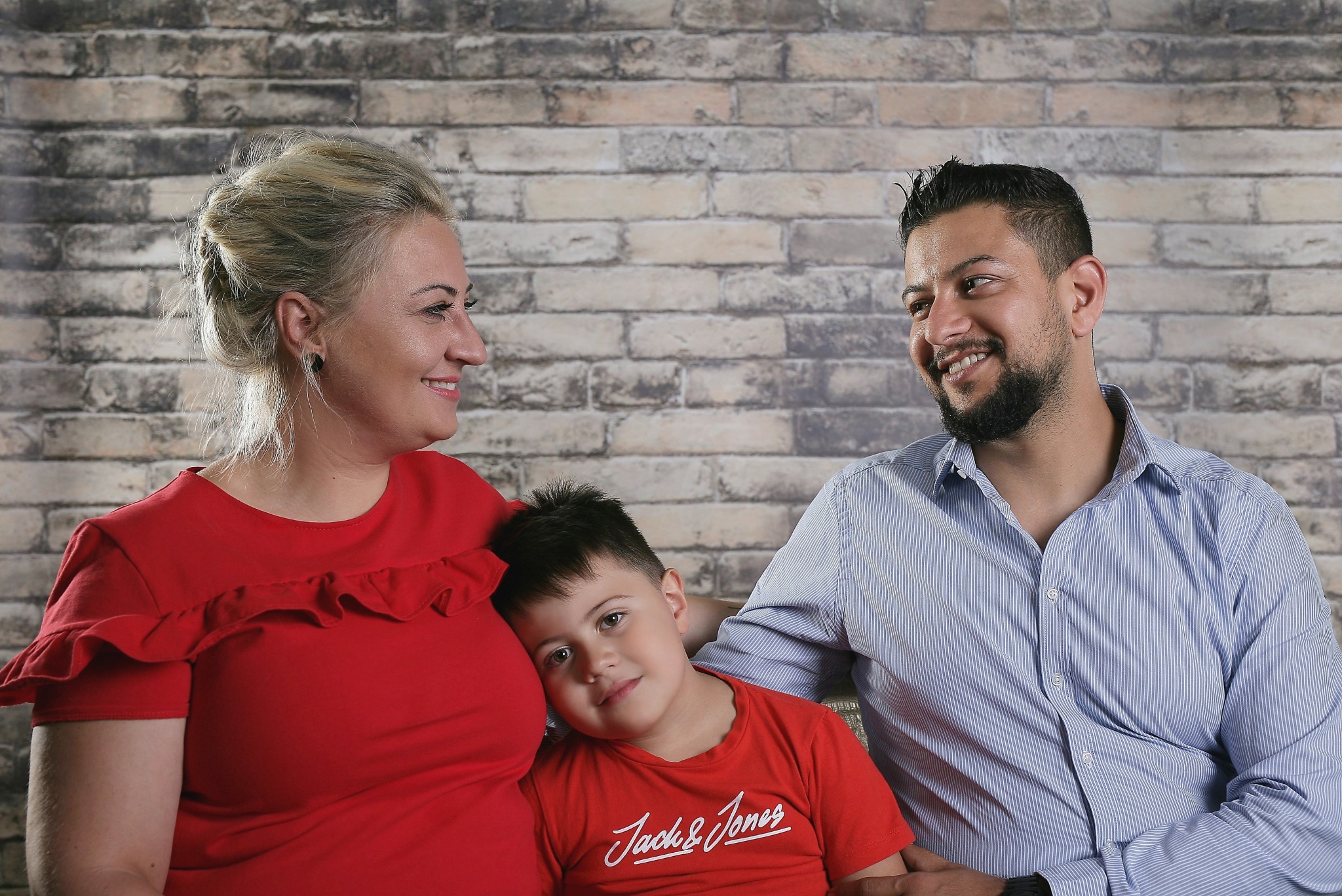 Uśmiechnięta rodzina: matka w czerwonej bluzce z falbankami, ojciec w błękitnej koszuli w paski i syn w czerwonej koszulce Jack&Jones, pozują na tle ściany z cegły.