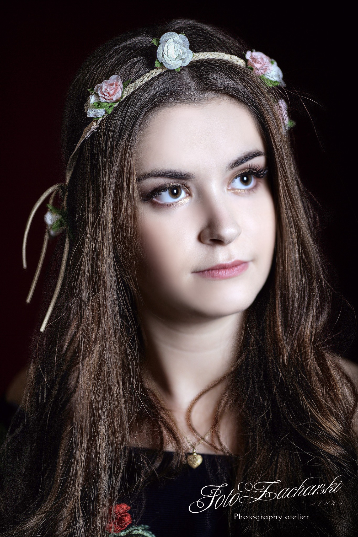 Portret młodej kobiety z wiankiem z białych i różowych kwiatów na głowie, z długimi brązowymi włosami i delikatnym makijażem, spoglądającej w górę na ciemnym tle.