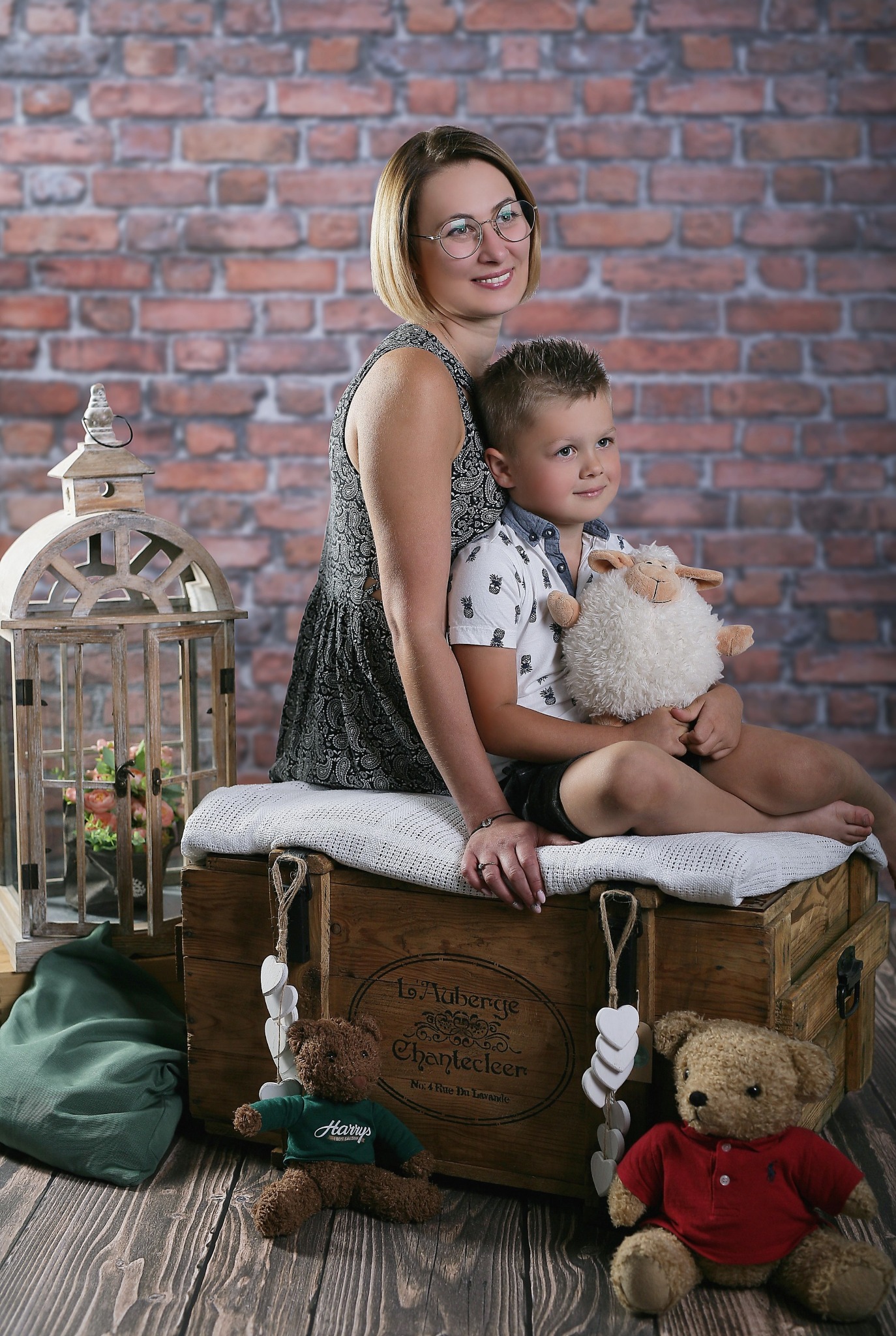 Kobieta w okularach i chłopiec z pluszową owcą siedzą na drewnianej skrzyni z dekoracjami, tło z cegieł