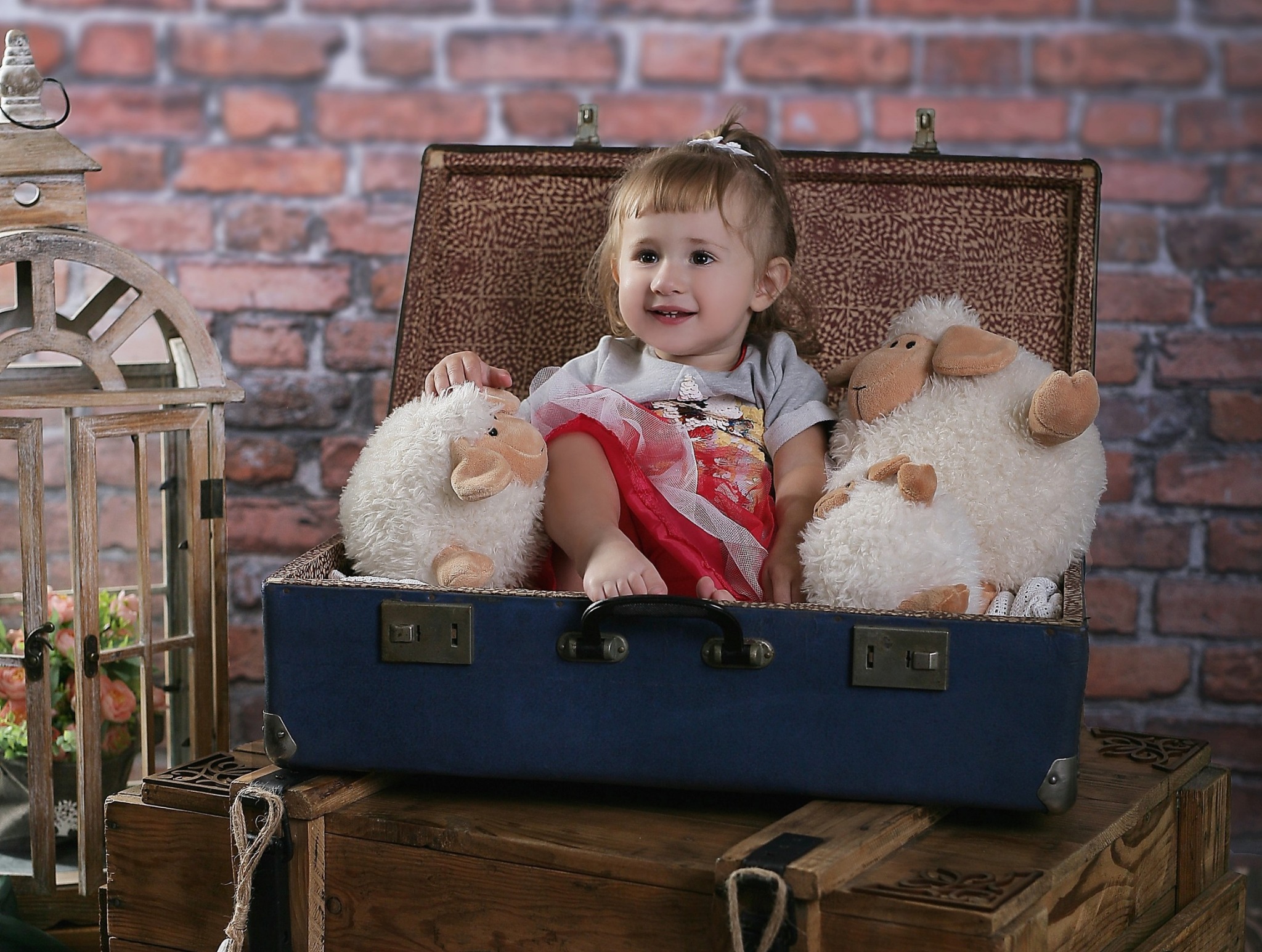 Uśmiechnięta dziewczynka w sukience siedzi w niebieskiej walizce otoczonej pluszowymi owieczkami, na tle ceglanej ściany i drewnianej skrzynki, podczas sesji dziecięcej w Nowym Sączu.
