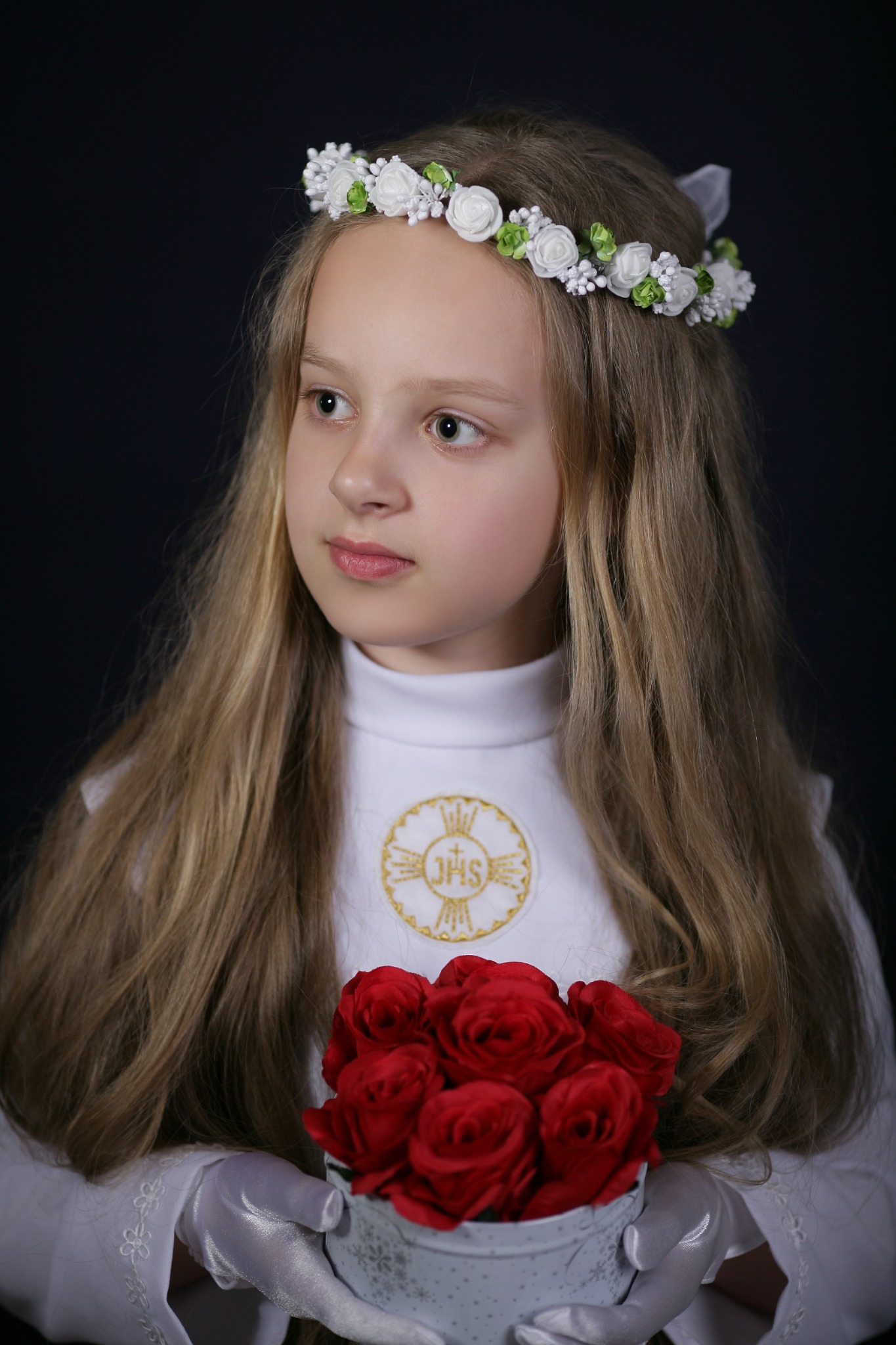Portret dziewczynki w stroju komunijnym z wiankiem z białych kwiatów i bukietem czerwonych róż, na ciemnym tle.