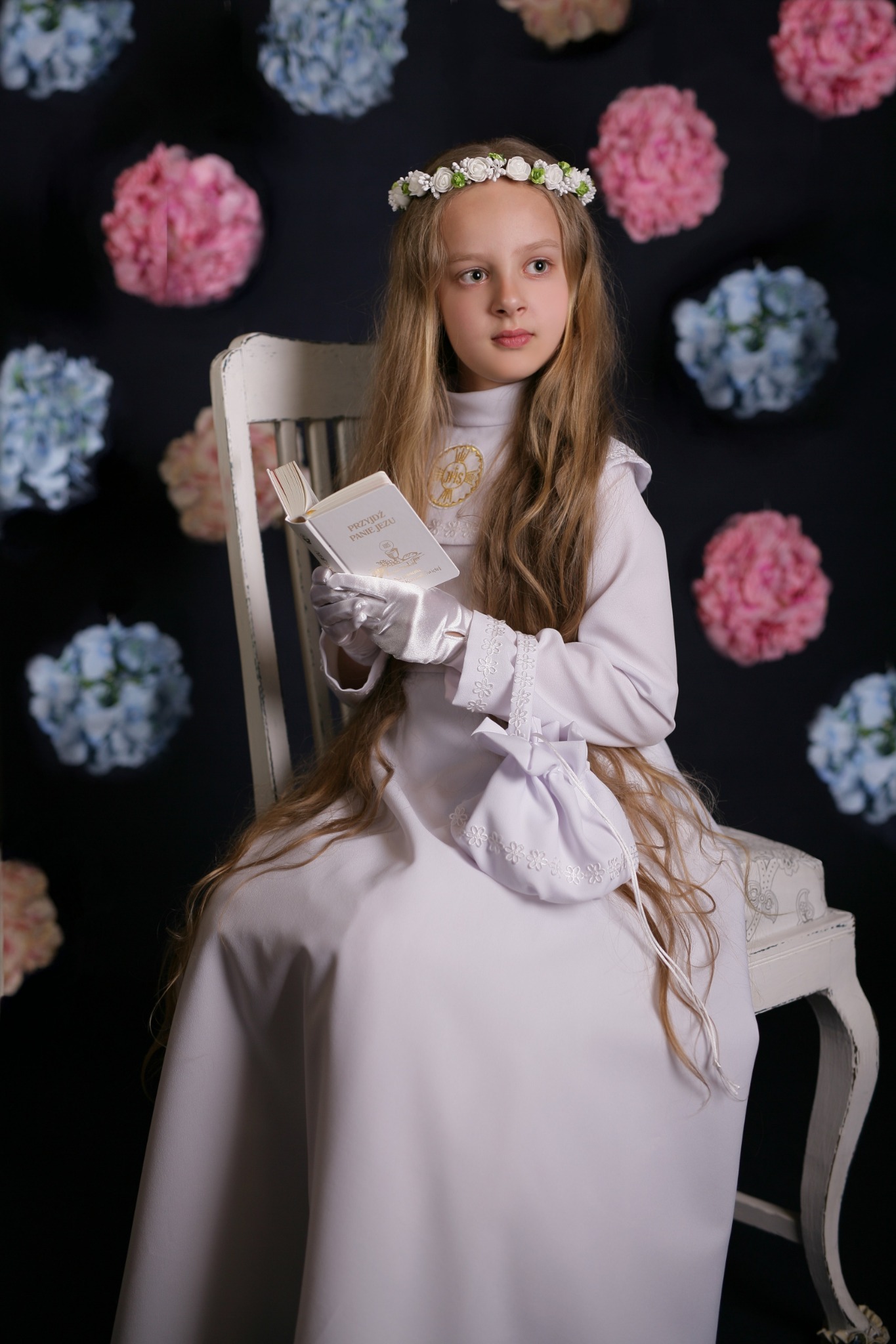 Dziewczynka w białej albie, z wiankiem na głowie, siedzi na krześle, trzymając książeczkę do Pierwszej Komunii Świętej; tło stanowią kwiaty w odcieniach różu i błękitu.