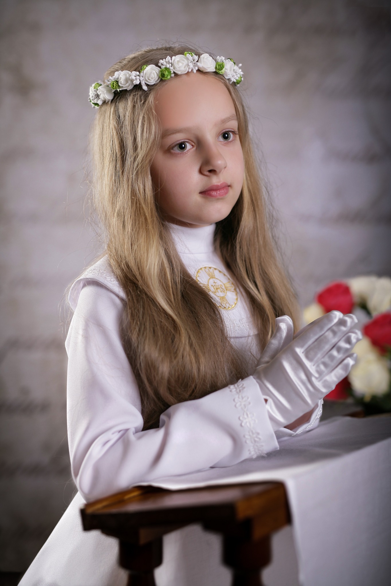 Dziewczynka w białej sukience komunijnej z wiankiem na głowie i rękawiczkami, z rękami złożonymi do modlitwy, siedząca przy stole z bukietem róż w tle.