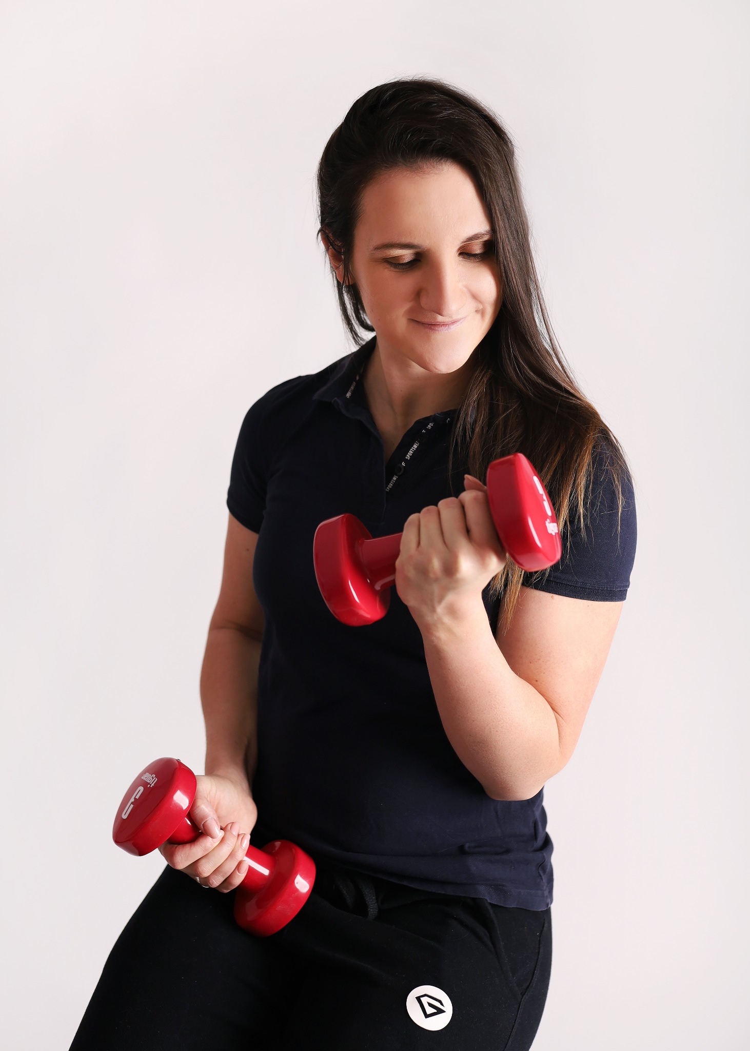 Kobieta w czarnym stroju sportowym ćwiczy biceps z czerwonymi hantlami na białym tle, spoglądając w dół na unoszoną rękę.