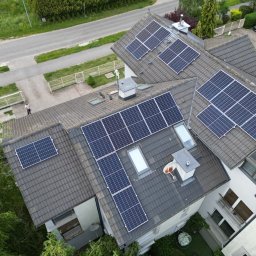 COSINUS fotowoltaika - Widok z góry na dom z panelami słonecznymi na dachu; widoczny fragment ulicy, trawnik i osoba stojąca obok ogrodzenia.