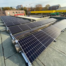 COSINUS fotowoltaika - Instalacja paneli słonecznych na płaskim dachu z widocznymi elementami montażowymi i fragmentem sylwetki instalatora w prawym górnym rogu, w tle budynki miejskie i stacja benzynowa.