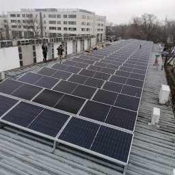 COSINUS fotowoltaika - Instalacja paneli słonecznych na falistym dachu z blachy, widoczni dwaj instalatorzy w oddali, budynek mieszkalny w tle.