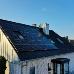 Instalacja fotowoltaiczna na dachu domu jednorodzinnego z białej cegły, widoczne okna dachowe, komin i antena satelitarna, w tle błękitne niebo.