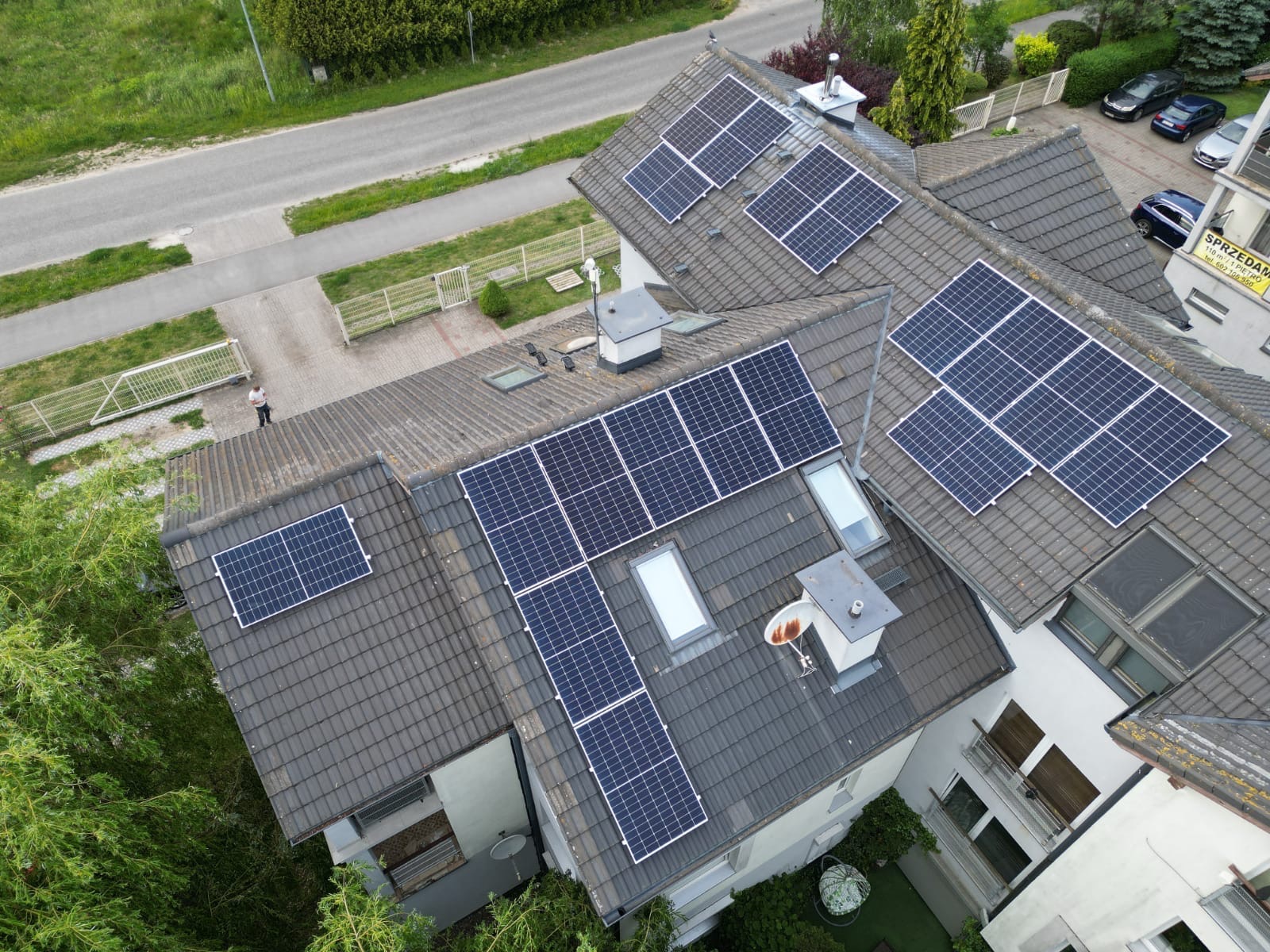 Widok z góry na dom z panelami słonecznymi na dachu; widoczny fragment ulicy, trawnik i osoba stojąca obok ogrodzenia.