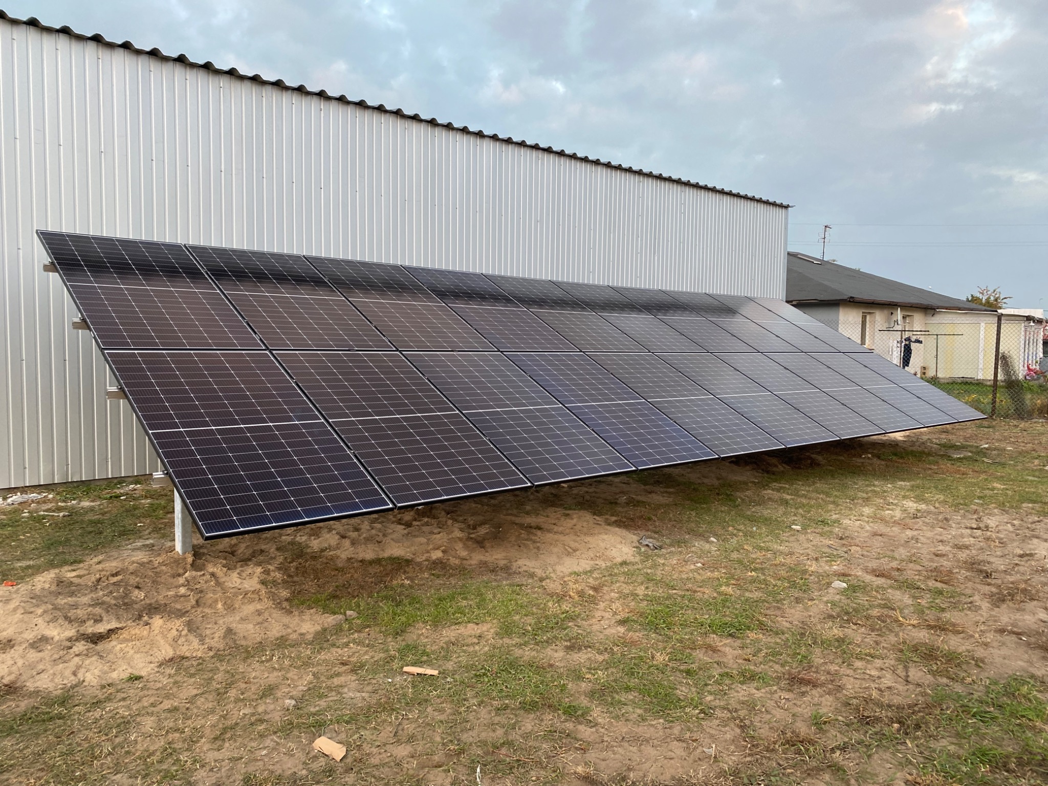 Rząd paneli słonecznych ustawionych na ziemi, widoczne metalowe podpory, w tle budynek z blachy falistej i fragment domu.