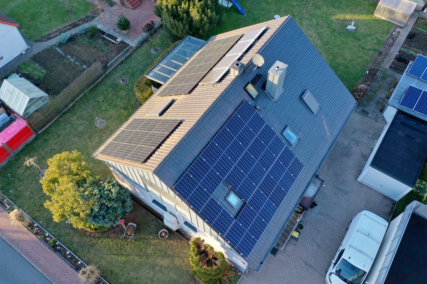 Widok z lotu ptaka na dom jednorodzinny z panelami słonecznymi na dachu, obok garażu zaparkowany biały bus, zadbany ogród z warzywnikiem i szklarnią.