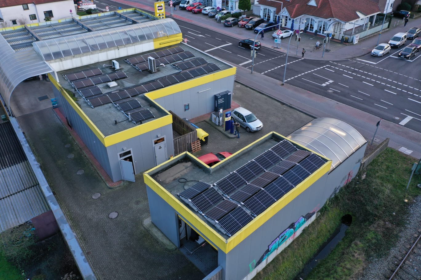 Widok z góry na budynki z panelami fotowoltaicznymi na dachach, stacja benzynowa z dystrybutorem, zaparkowany samochód osobowy i fragment skrzyżowania ulic.