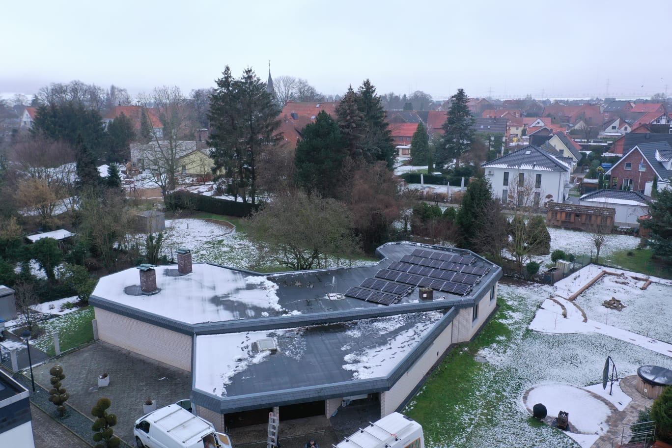 Widok z góry na dom z panelami słonecznymi na dachu, częściowo pokrytym śniegiem, z dwoma białymi furgonetkami zaparkowanymi przed garażem i okoliczną zabudową w tle.
