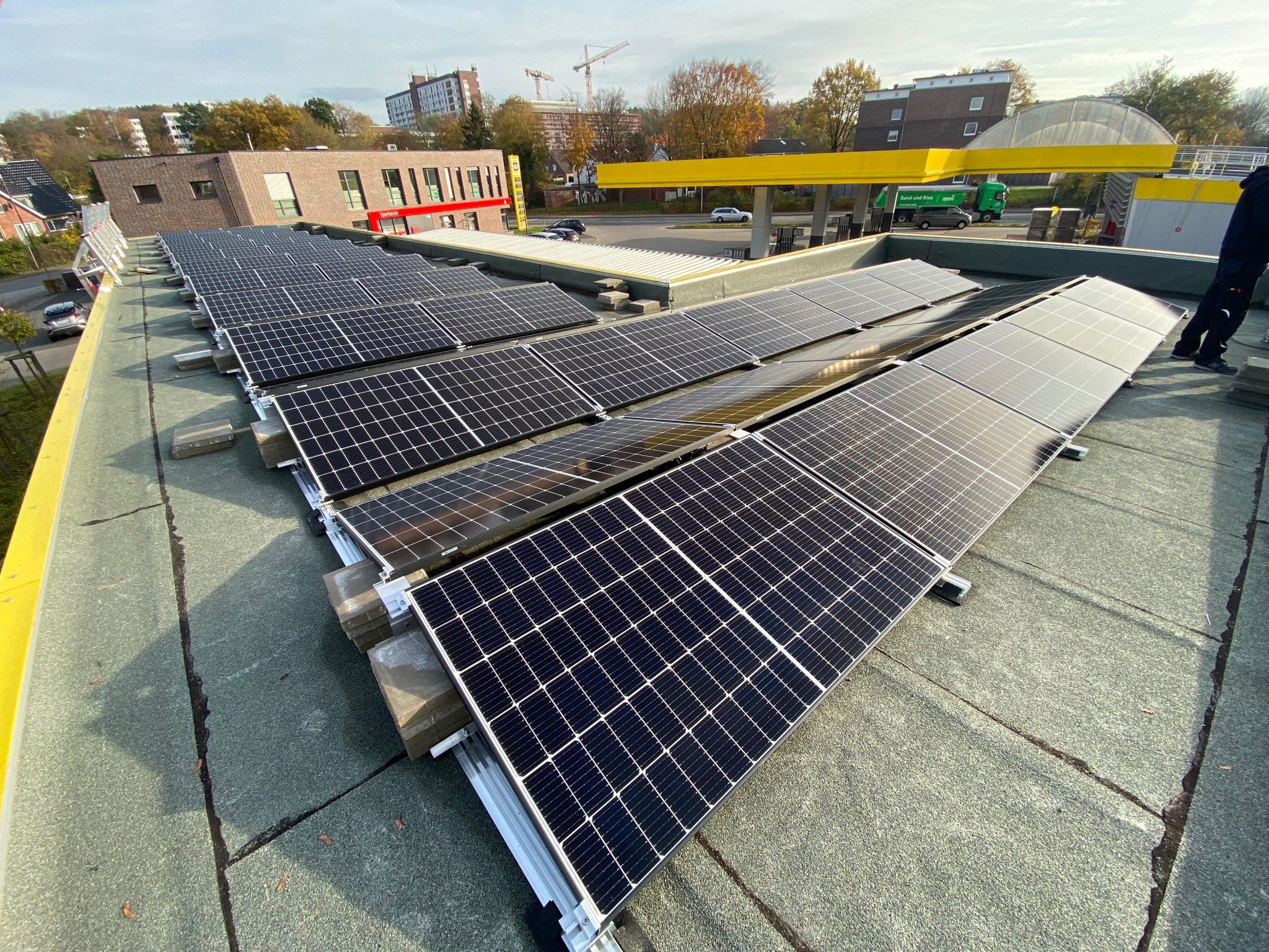 Instalacja paneli słonecznych na płaskim dachu z widocznymi elementami montażowymi i fragmentem sylwetki instalatora w prawym górnym rogu, w tle budynki miejskie i stacja benzynowa.